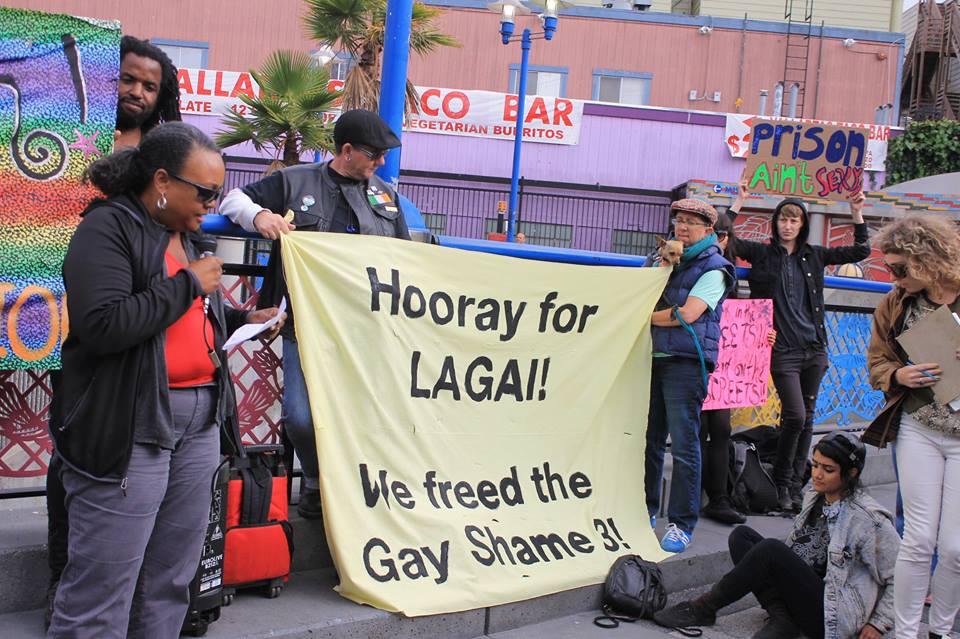 folks gathered at the 16th street bart station for a press conference holding a banner that reads: "hooray for lagai! we freed the gay shame 3!" while someone stands in the background holding a sign reading "prison ain't sexy"
