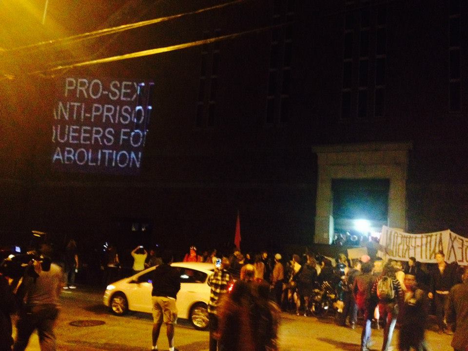 a crowd with banners gather on the north side of the armory building with a projection that reads: "pro-sex anti-prison queers for abolition"