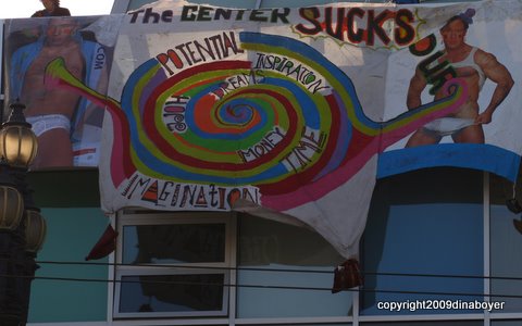 Photo of the banner drop at the De-Center the Center action! With two beefcakes in tightie-whities flanking a message saying "The Center Sucks" and a colorful whirly circle with the words "Potential, Inspiration, Hope, Dreams, Money, Time, Imagination" which are all things that the Center is sucking like an energy vortex.