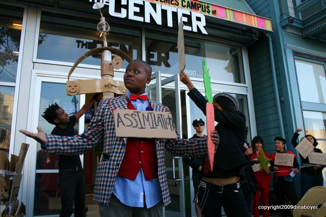 Image of someone wearing business attire that would befit a dandy living off a sizeable nonprofit salary (the person wears a sign saying "Assimilation" and also holds a faux carrot); a cross is being erected in the background and some other action-goers bear signs alongside it in front of the main doors to the LGBT Center in San Francisco.