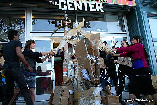 people gathered around a pile of cardboard props in front of the locked-open main entrance of the lgbt center at 1800 market street, two people cross cardboard swords before competitively striking to break a large cardboard "stone" with the cardboard polarity meter in the background