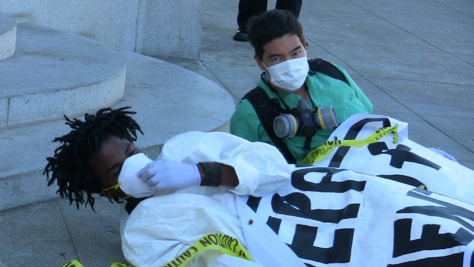 two people laying on the ground apparently ill at the steps of the department of public health under a blanket with respirator face masks and yellow tape