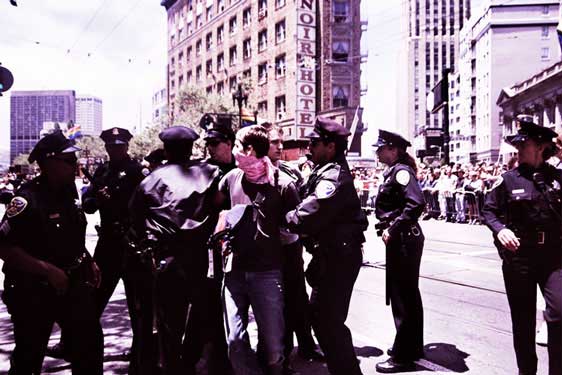  person in pink bandana being mobbed by a large group of police inside the market street gay pride parade route
