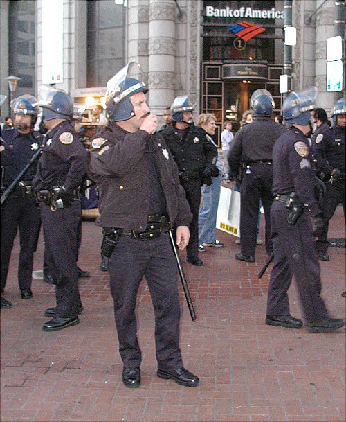 riot police milling around in front of the old bank of america location at the powell turnaround with the plastic face shields of their riot helmets up and long batons 