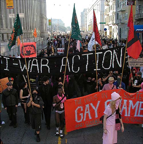 an overhead shot of hundreds of people marching down mcallister from san francisco city hall between larkin toward hyde under the massive black "anti-war action" banner and the red "gay shame" banner in front with a few people in pink