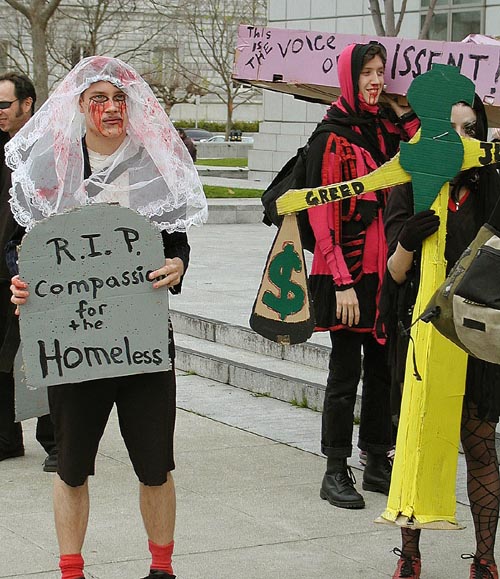 people dressed as bloodstained mourners with blood and lace, one holding a coffin labeled "the voice of dissent", another with a cardboard tombstone reading "r.i.p. compassion for the homeless"