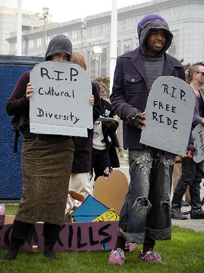two mourners carrying cardboard tombstones reading "r.i.p. cultural diversity" and "r.i.p. free ride" respectively