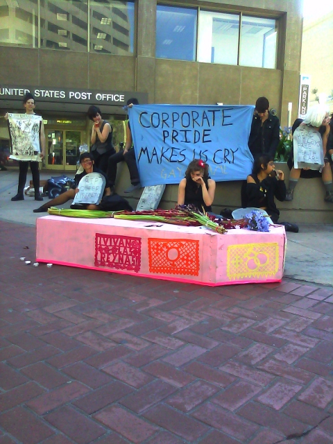 people gathered at the planter in the area in front of the post office at market and hayes with a pink cardboard coffin plastered with multicolored queer papel picada and wilted violet hyacinth and magenta gladioli on the lid holding a blue "corporate pride makes us cry" banner
