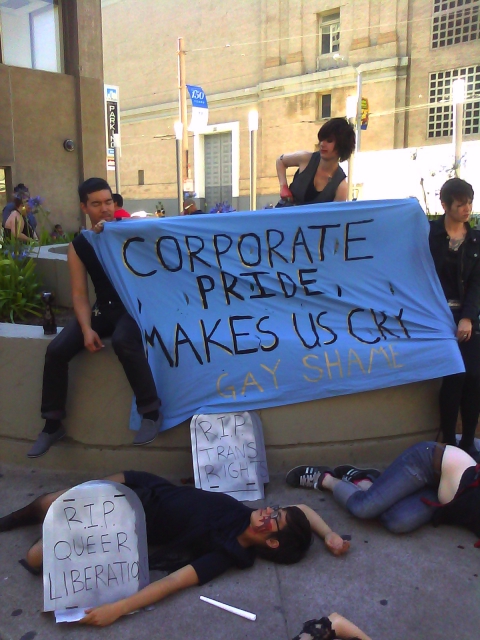 people holding a blue banner that reads "corporate pride makes us sick" sitting on the planter in front of the post office at market and hayes and some other people apparently playing dead on the ground with cardboard tombstones reading "r.i.p. queer liberation" and "r.i.p. trans rights"