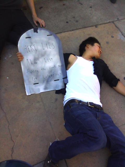a person apparently playing dead on the ground holding a cardboard tombstone reading "here lies those lost fighting the good fight for liberation"