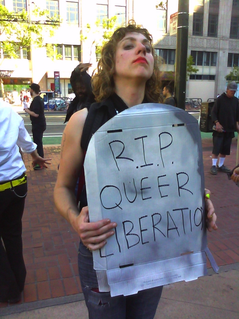 person holding a cardboard tombstone that says "r.i.p. queer liberation"
