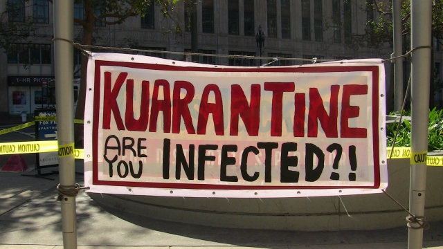 a banner attached to the flagpoles in front of the old post office at market and hayes reading: "kuarantine are you infected?"