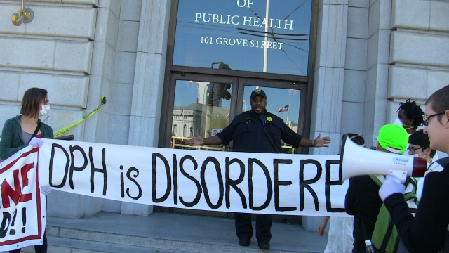 a police officer on the steps of the department of public health at grove and polk blocking a banner that says "dph is disordered"