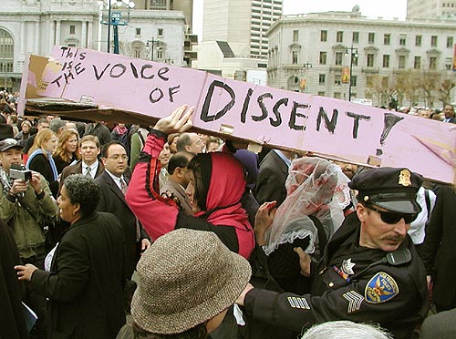 people carrying a pink coffin that reads "this is the voice of dissent" through a crowd at newsom's inauguration ceremony at civic center plaza