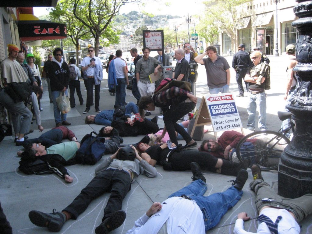 a large group of people stage a die-in on market street on the sidewalk in front of the new coldwell banker location 