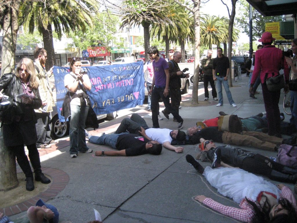 a larger group of people staging a die-in on market street near castro in front of the new coldwell banker location