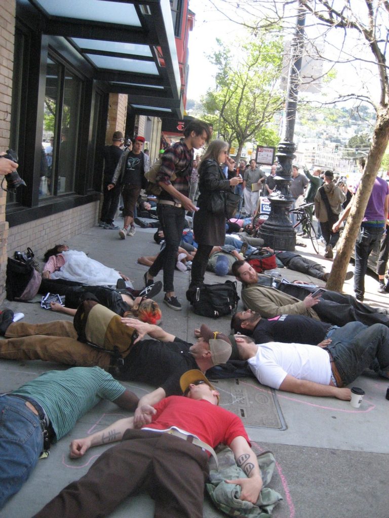 a large people staging a die-in on the sidewalk in front of the new market street coldwell banker location near castro