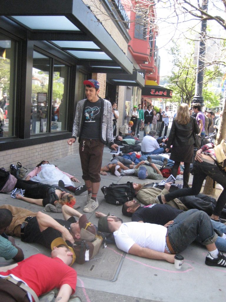 a large people are staging a die-in on the sidewalk in front of the coldwell banker location on market near castro