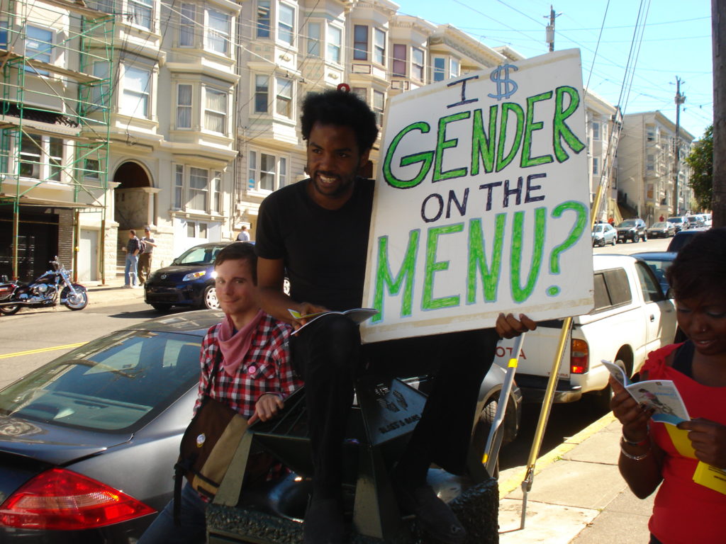 a person holding a sign that says "is gender on the menu" while sitting on top of a trashcan at the corner of 19th and castro on the sidewalk in front of the HRC store's original location