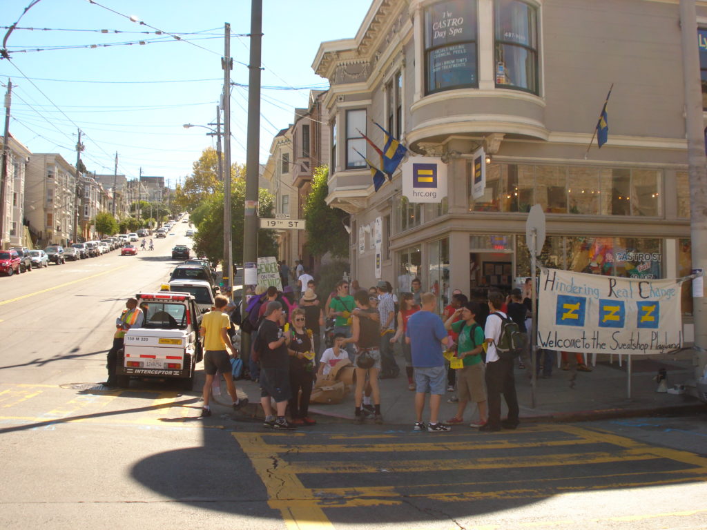 a group of people with signs and props gathered on the sidewalk at the corner of 19th and castro in front of the original hrc.org store location