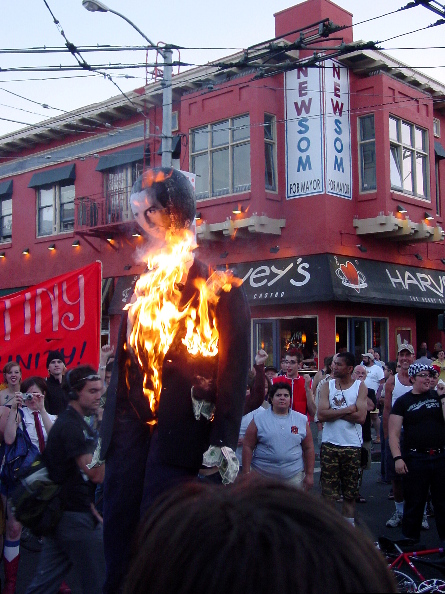 people gather around watching the gavin newsom effigy now fully ablaze as they hold the intersection of 18th and castro directly in front of a giant "newsom for mayor" sign above harvey's restaurant. 