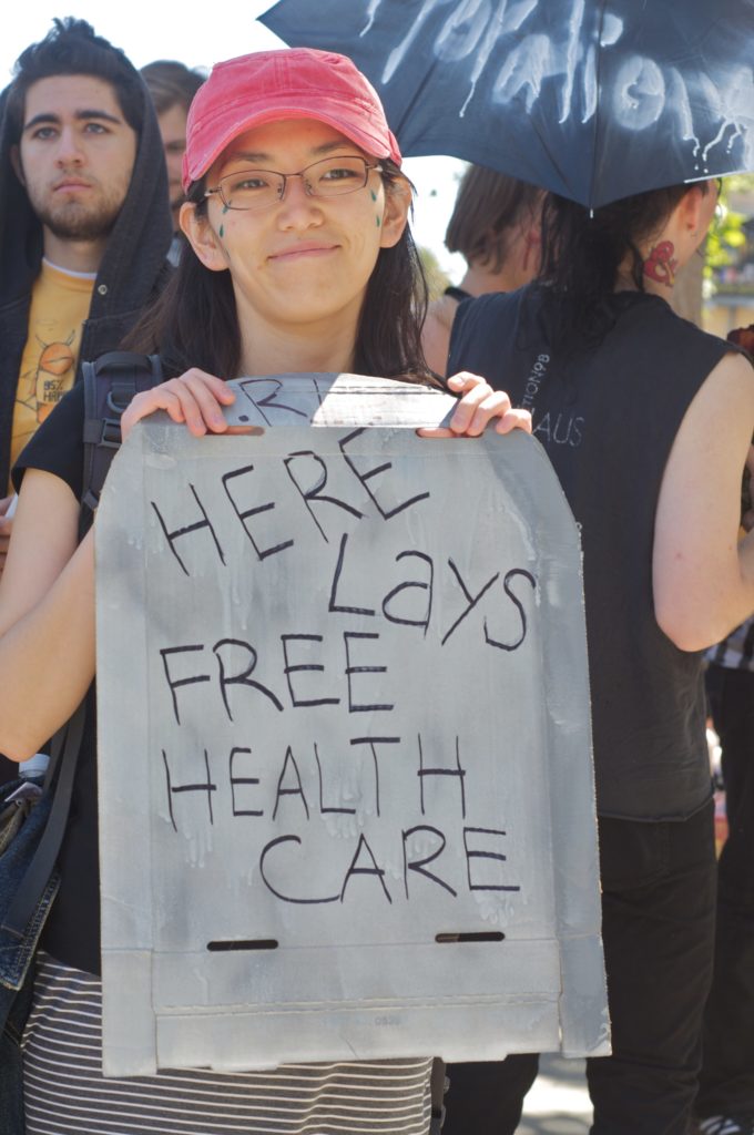person holding a cardboard tombstone reading "here lays free health care"