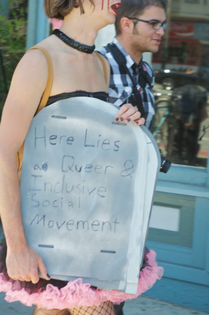 person carrying a cardboard tombstone that reads "here lies a queer & inclusive social movement"