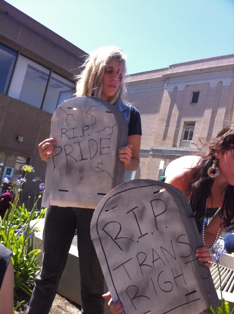 people holding cardboard tombstones reading "r.i.p. pride" and "r.i.p. trans rights" gathered on the large concrete planter in the post office area at market and hayes