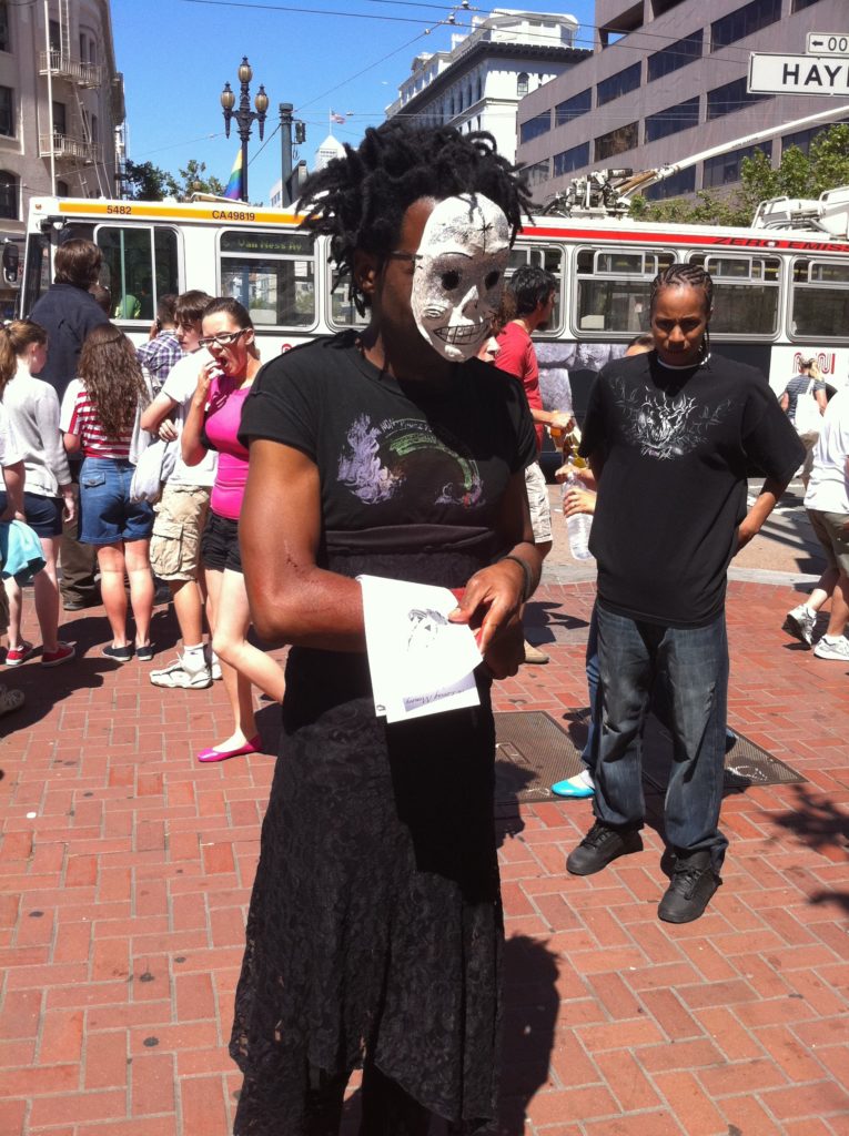 person in a sugar skull mask and a black lace skirt handing out flyers at market and hayes