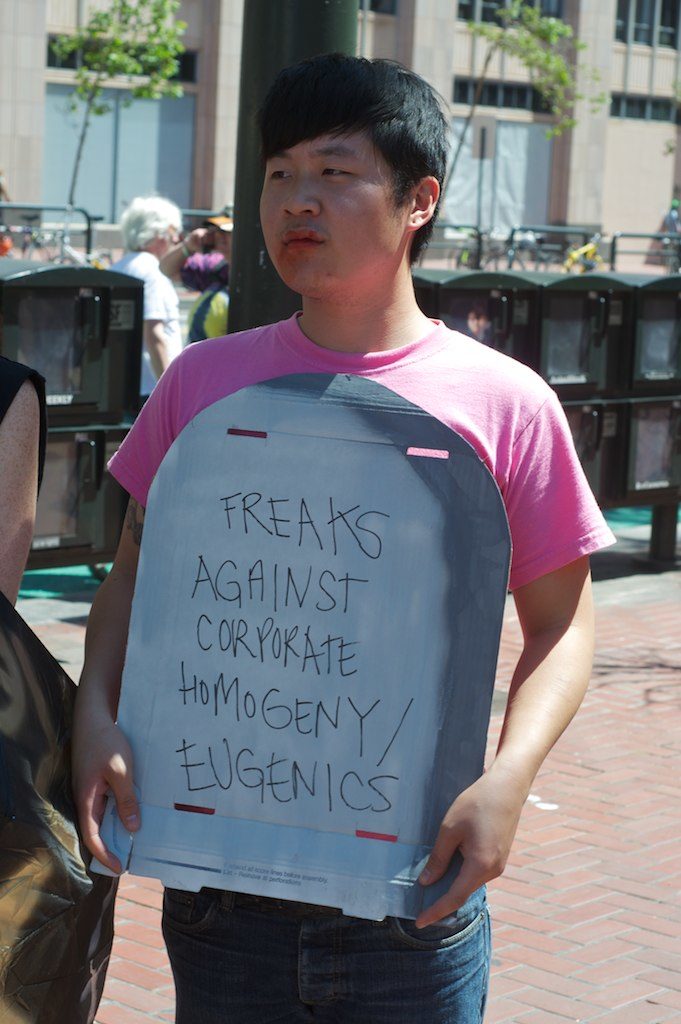 person holding a cardboard tombstone that reads "freaks against corporate homogeneity / eugenics"