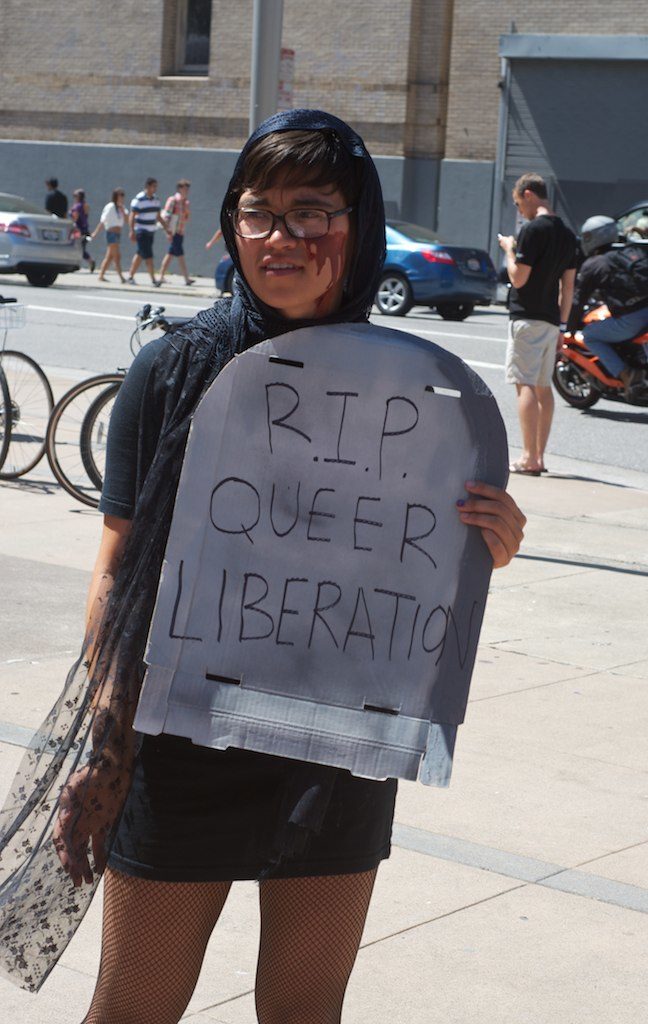 person holding a cardboard tombstone that reads "r.i.p. queer liberation" in the sidewalk area in front of the post office at market and hayes