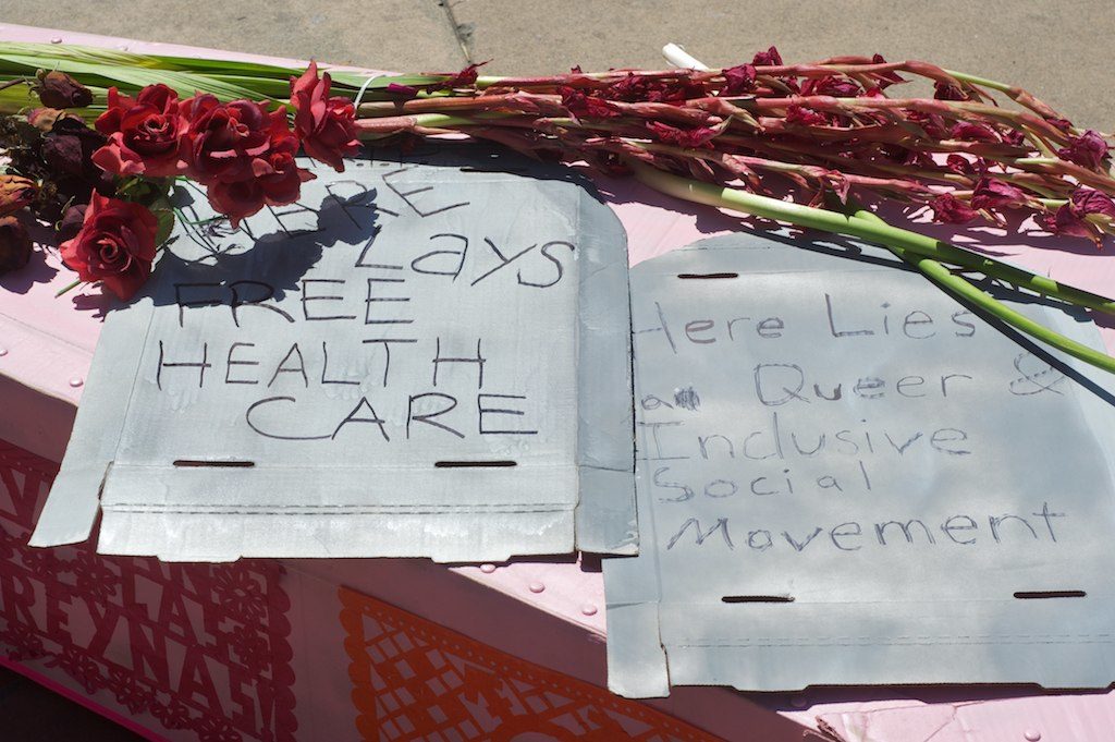close up of indehiscent pink gladiolis and roses and cardboard tombstones on the lid of the pink cardboard coffin that read: "here lays free health care" and "here lies a queer & inclusive social movement" and papel picados plastered on the side of the coffin
