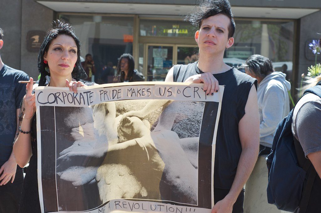 two people with apparently dour expressions holding a sign that says "corporate pride makes us cry"