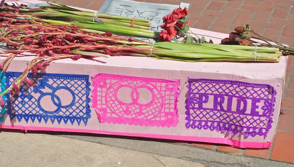 detail of a pink cardboard coffin covered with slightly wilting indehiscent pink silk stemmed gladioli and multicolored queer papel picado pasted on the side