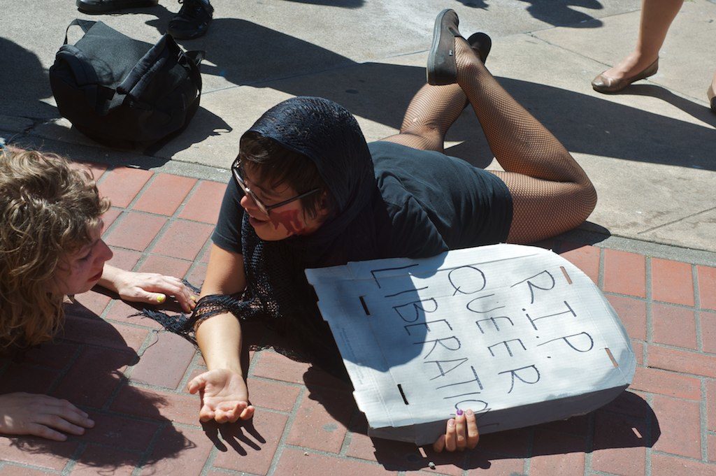 people crawling on the ground sobbing, one holding a cardboard tombstone that reads "r.i.p." queer liberation