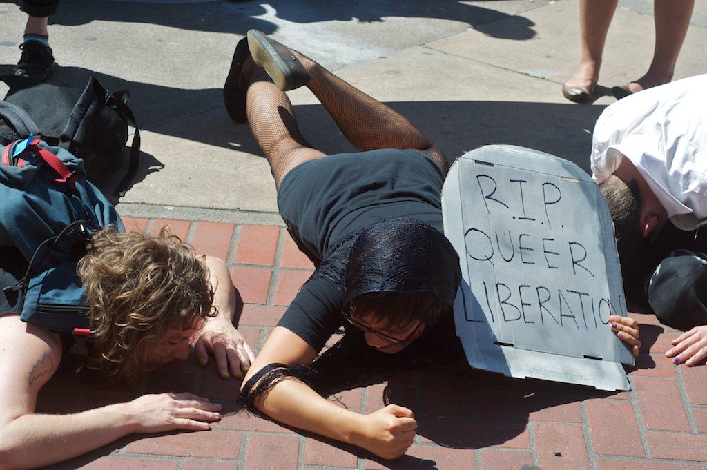 people on the ground wailing, one person holding a cardboard tombstone that reads "r.i.p. queer liberation"