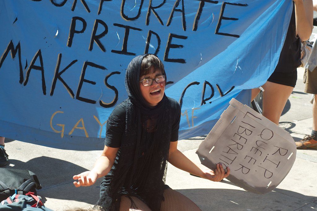 person wailing next to a baby blue banner that says "corporate pride makes us cry" and holding a cardboard tombstone that says "r.i.p. queer liberation"