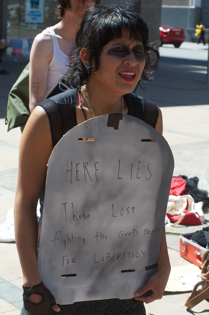 person sobbing holding a cardboard tombstone reading "here lies those lost fighting the good fight for liberation"