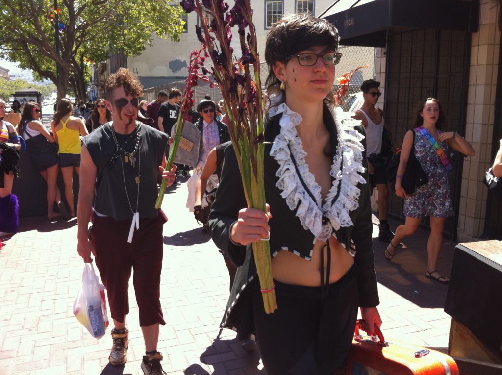 the procession continues with a person walking down market holding a few rubber-banded stems of wilted violet hyacinth
