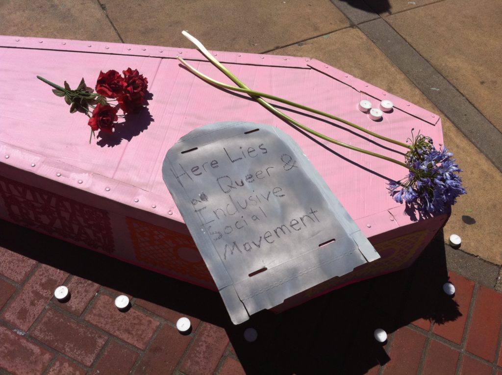 tealight candles dot a nosegay of red roses and two stems of violet hyacinth and a cardboard tombstone reading "here lies a queer & inclusive social movement" on the lid of a pink cardboard casket