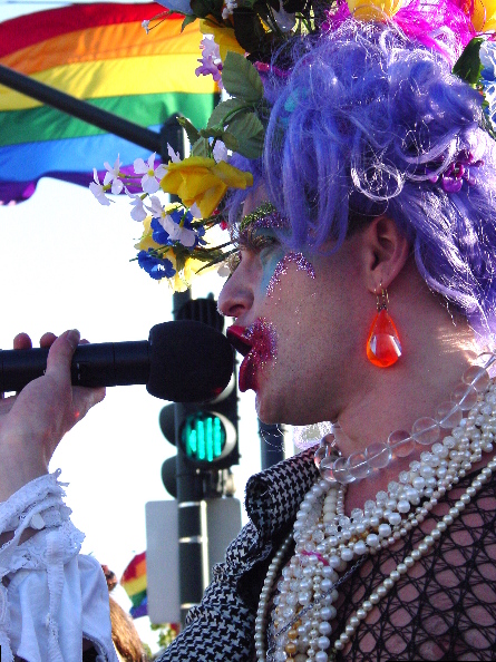 close up of a speaker with a microphone providing detail of their elaborate clothes, make-up, wig with flowers and jewelry. the pride flag looms in the background