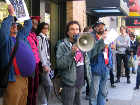 a person with a bullhorn with a group of people rallying on the sidewalk in front of the coldwell banker office on market near castro