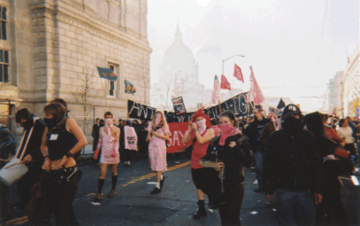 view of a few people in pink marching down mcallister in of some more people dressed in black marching in front of a large black "anti-war action" banner peeking behind signs and flags with the san francisco city hall dome looming in foggy silhouette