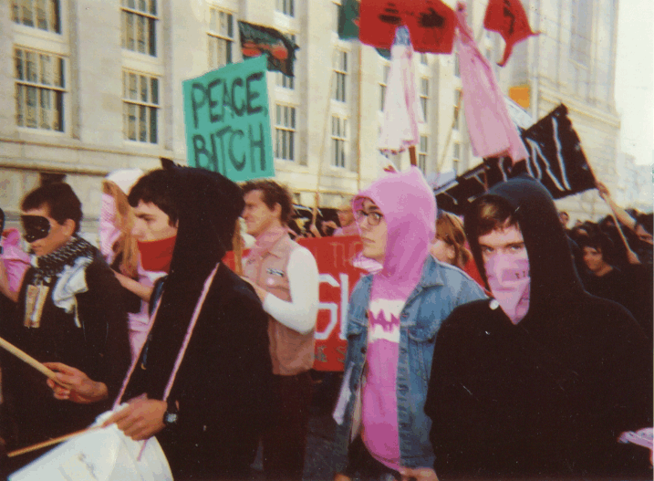 a group of people marching down mcallister from city hall between larkin and hyde some in pink and some in black. there is a prominent view of a turquoise sign that reads "peace bitch"