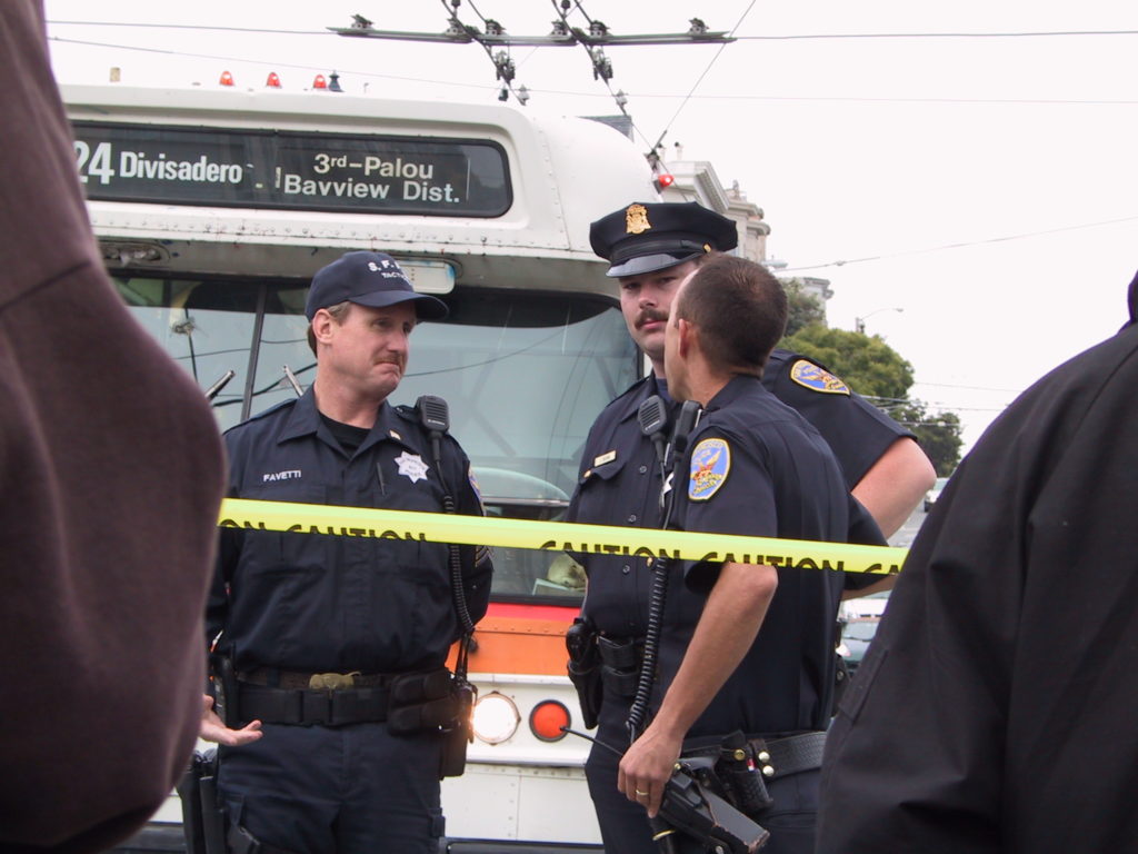 police officers stand around looking perplexed in front of a blocked 24 divisadero muni bus at castro and market behind a line of yellow caution tape