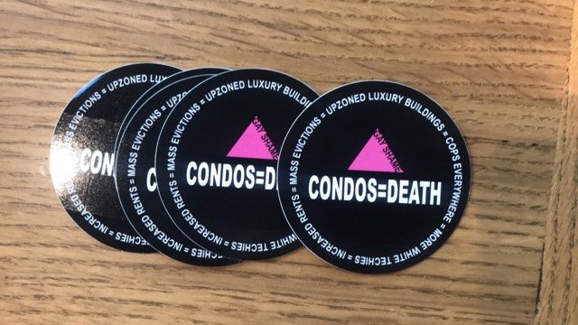black round stickers with an inverted pink triangle that says "GAY SHAME" and white text that reads: "CONDOS = DEATH" and white text that edges the circle's perimeter in an endless loop reading: "MASS EVICTIONS = UPZONED LUXURY BUILDINGS = COPS EVERYWHERE = MORE WHITE TECHIES = INCREASED RENTS = MASS EVICTIONS..."