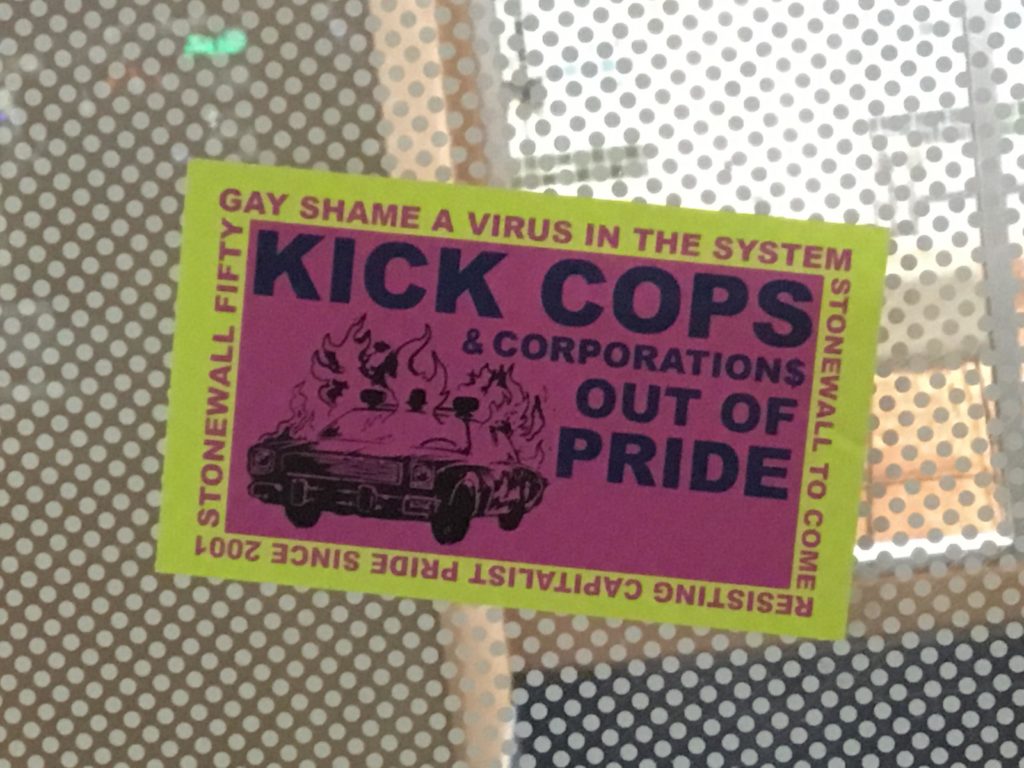 a pink and yellow green sticker with a police car on fire that reads "KICK COPS OUT AND CORPORATIONS OUT OF PRIDE" and written around the border "GAY SHAME A VIRUS IN THE SYSTEM" "STONEWALL FIFTY" "STONEWALL TO COME" "RESISTING CAPITALIST PRIDE SINCE 2001"