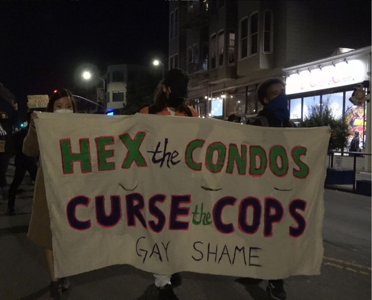 hand painted banner being held by three people leading a march down valencia in the evening which reads: hex the condos curse the cops gay shame