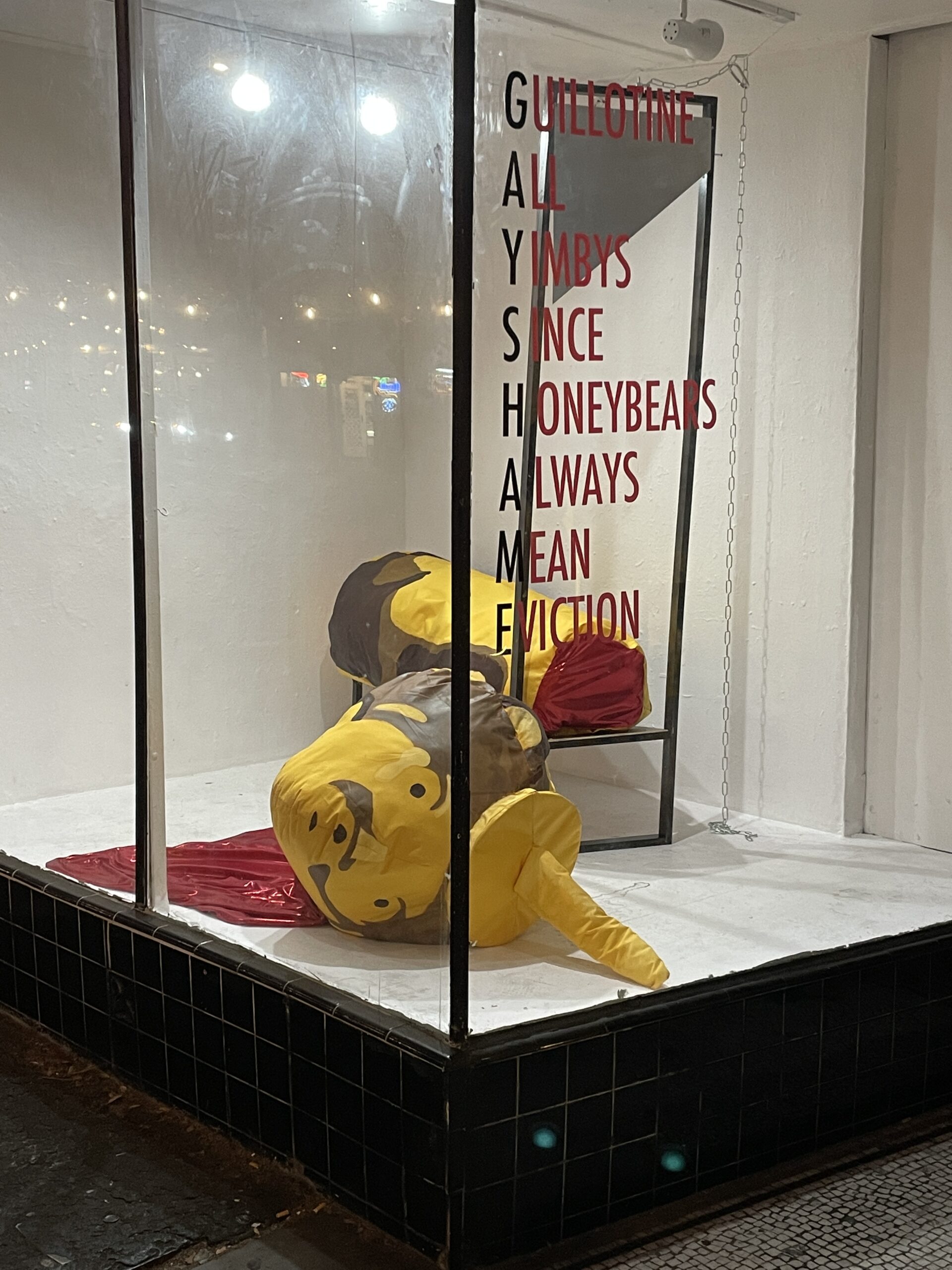 Photography of a honey bear that has been beheaded by a guillotine in a window display. On the window text reads "GUILLOTINE ALL YIMBYS SINCE HONEYBEARS ALWAYS MEAN EVICTION"
